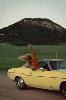 A woman leaning out of a yellow car's window, hair flying, on a country road.