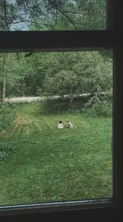Two small children sitting in a green yard, seen through a window.